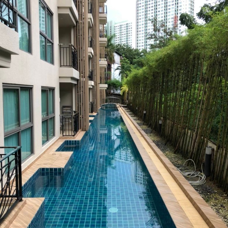 Narrow rectangular swimming pool with blue tiled deck between a beige apartment building and a bamboo fence with high rise buildings in the distance