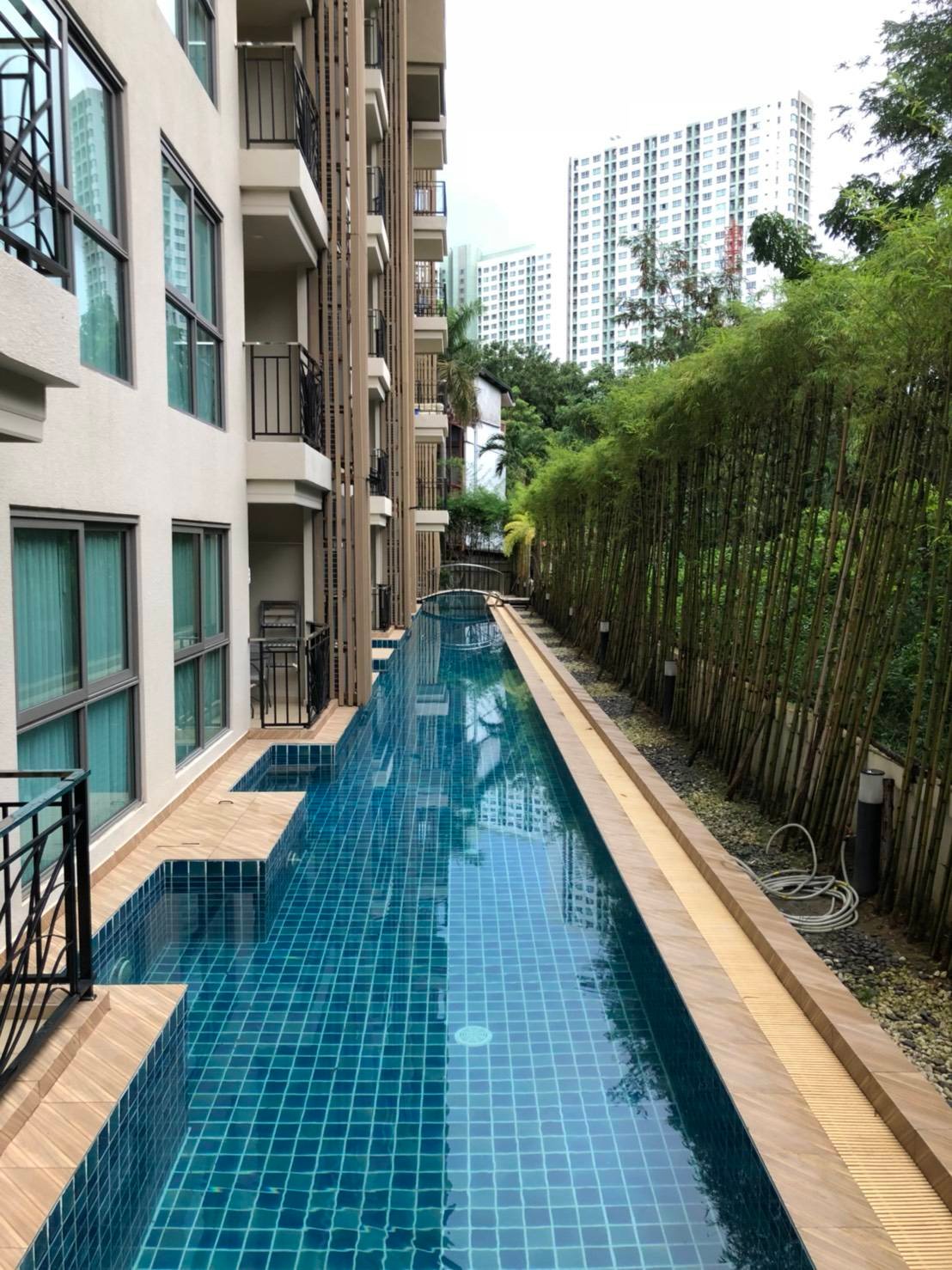 Narrow rectangular swimming pool with blue-tiled deck between a beige apartment building and a bamboo fence, with high-rise buildings in the distance.