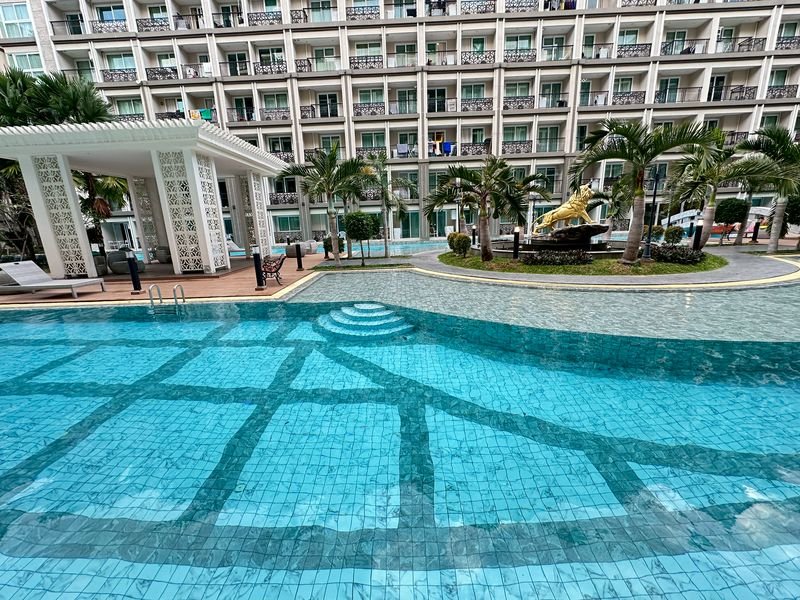 Hotel resort pool with turquoise tiles, surrounding palm trees, and a multi-story building with balconies in the background.