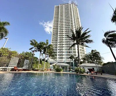 Tall white high-rise residential tower with balconies, seen behind a central swimming pool, palm trees and lounge chairs.