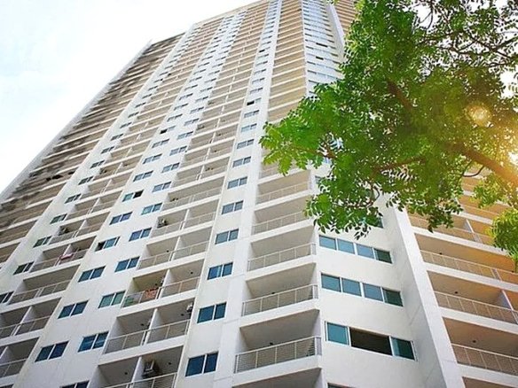 Tall white apartment building with many balconies viewed from below with a tree and sun flare on the right