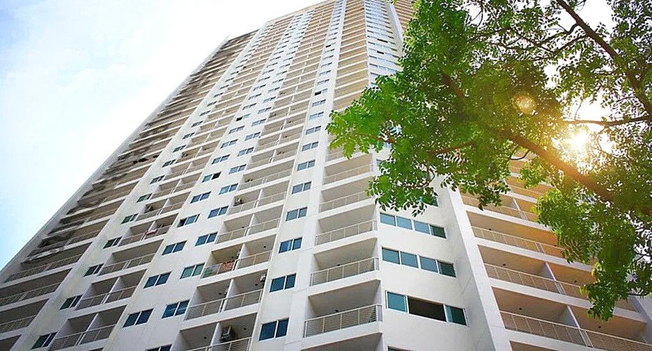 Tall white apartment building with many balconies, viewed from below, with a tree and sun flare on the right.