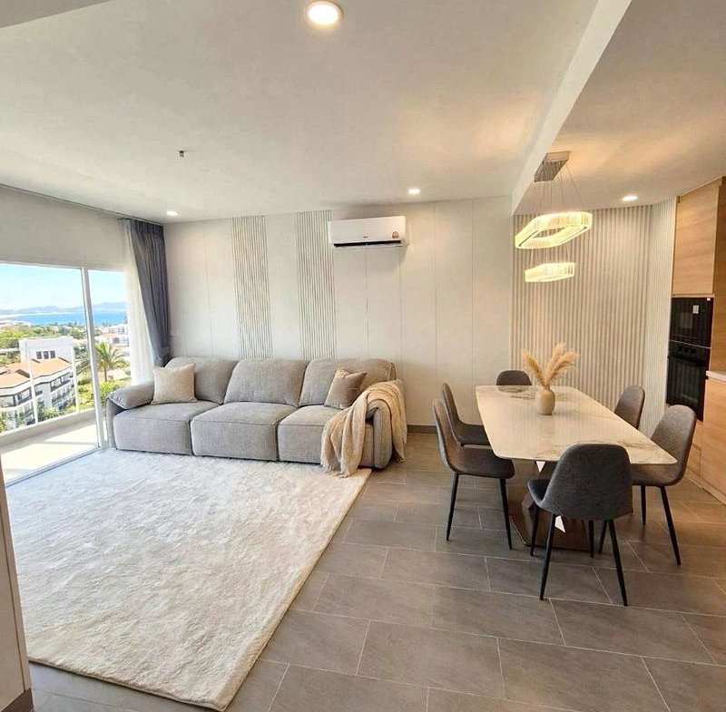 Open-plan living and dining area with a light gray sofa, cream rug, and a long dining table near a pastel kitchen island, balcony view in the background.