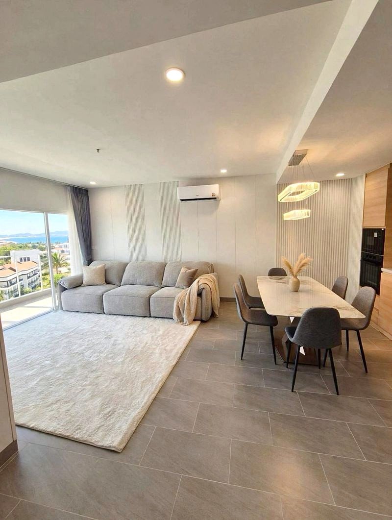 Open-plan living and dining area with a light gray sofa, cream rug, and a long dining table near a pastel kitchen island, balcony view in the background.