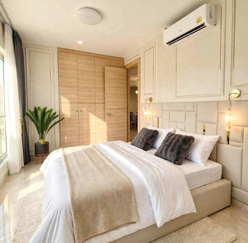 Bright, modern bedroom with a padded headboard bed, white linens, beige throw, two gray pillows, and a large wooden wardrobe nearby.