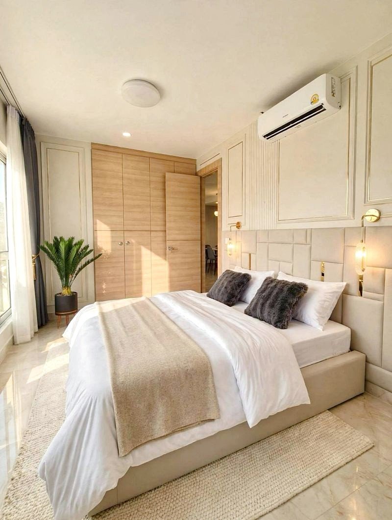 Bright, modern bedroom with a padded headboard bed, white linens, beige throw, two gray pillows, and a large wooden wardrobe nearby.
