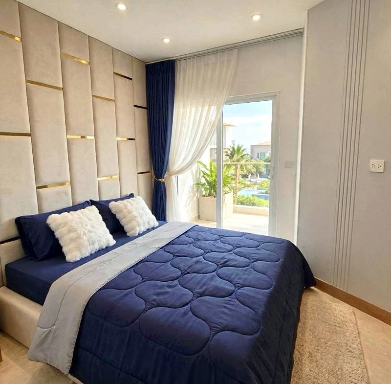 Bedroom with a navy quilted bed, white fluffy pillows, and a padded beige paneled wall; sheer and blue curtains beside a glass door opening to a sunny balcony.