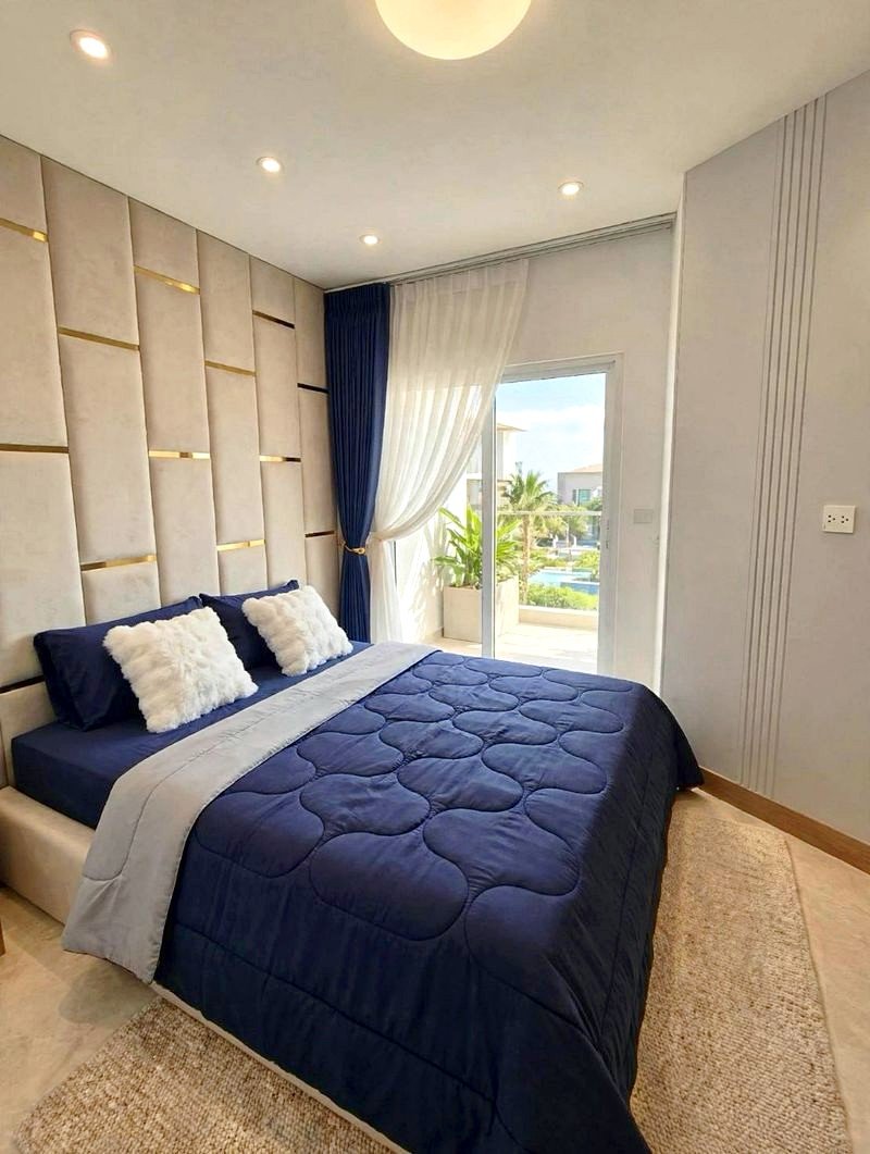 Bedroom with a navy quilted bed, white fluffy pillows, and a padded beige paneled wall; sheer and blue curtains beside a glass door opening to a sunny balcony.