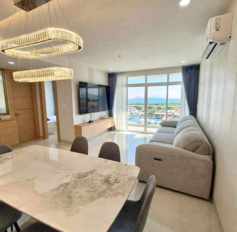 Open-plan living and dining area with a marble dining table, gray chairs, a crystal chandelier, and a gray sofa facing a wall TV.