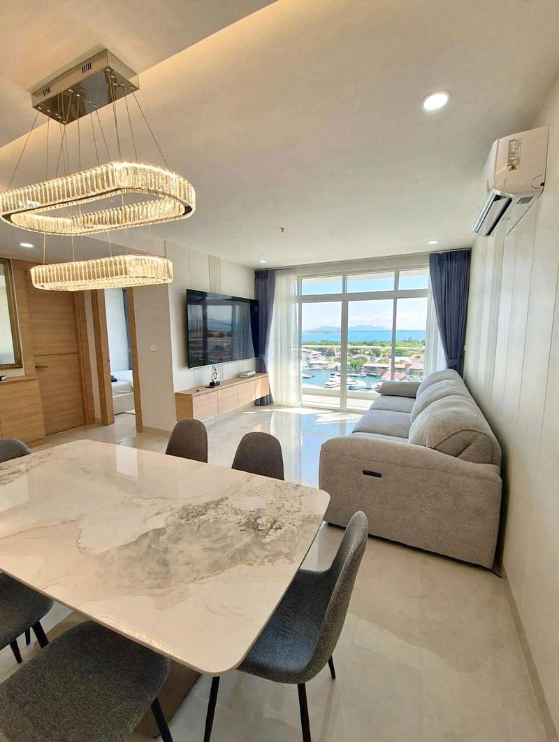 Open-plan living and dining area with a marble dining table, gray chairs, a crystal chandelier, and a gray sofa facing a wall TV.