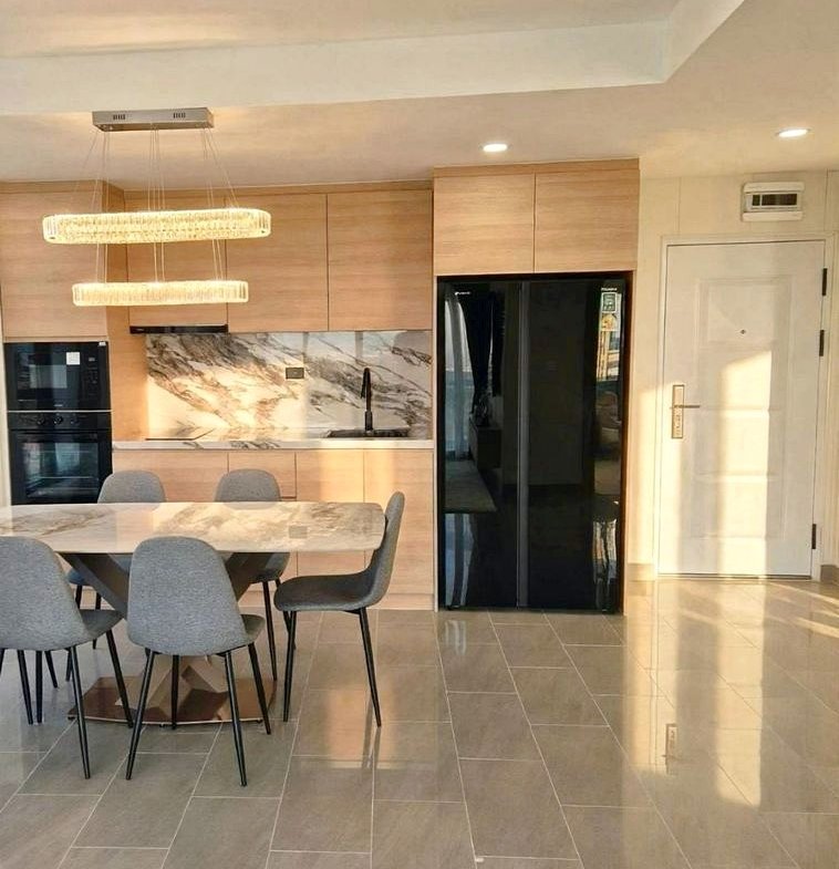 Open-concept kitchen and dining area with light wood cabinets, marble backsplash, pendant lights, and a marble dining table surrounded by gray chairs next to a large black refrigerator.
