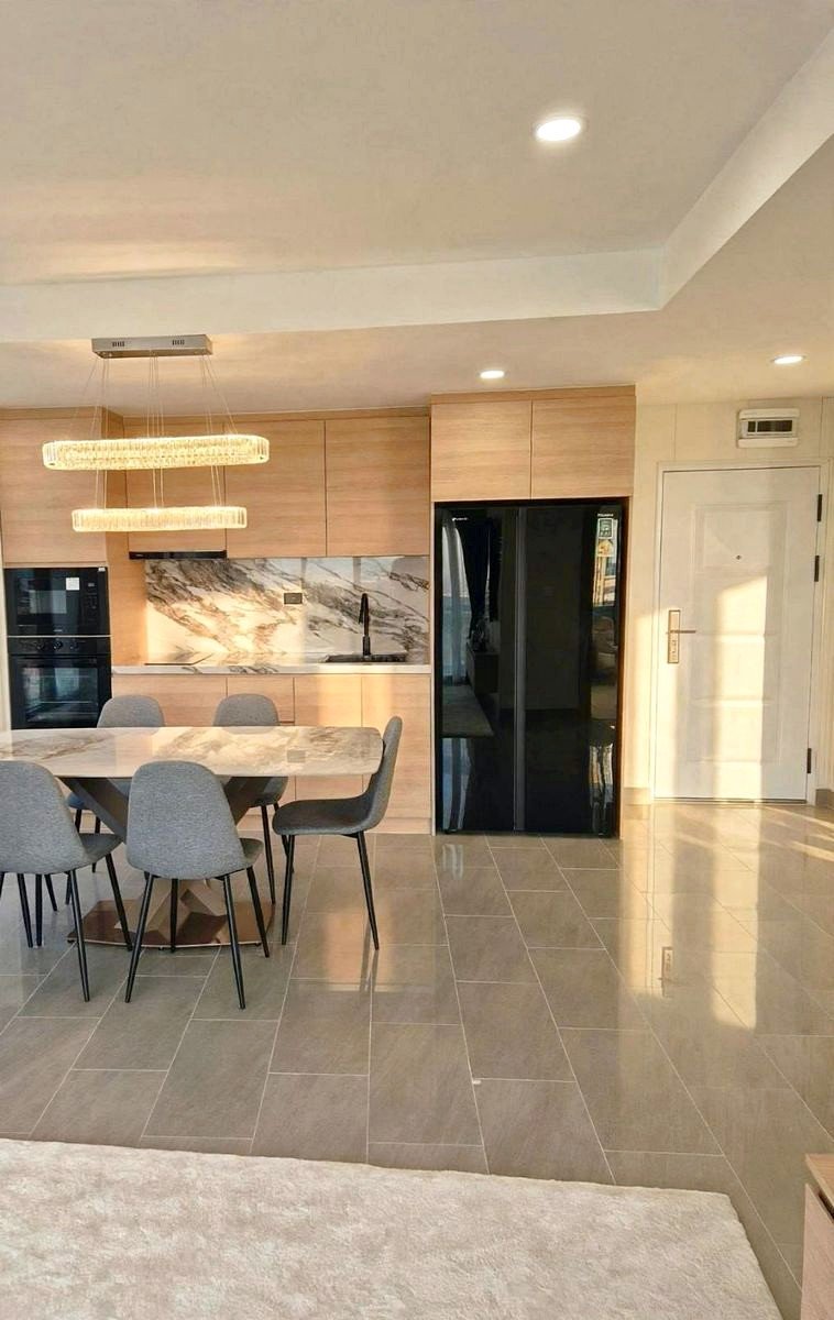 Open-concept kitchen and dining area with light wood cabinets, marble backsplash, pendant lights, and a marble dining table surrounded by gray chairs next to a large black refrigerator.