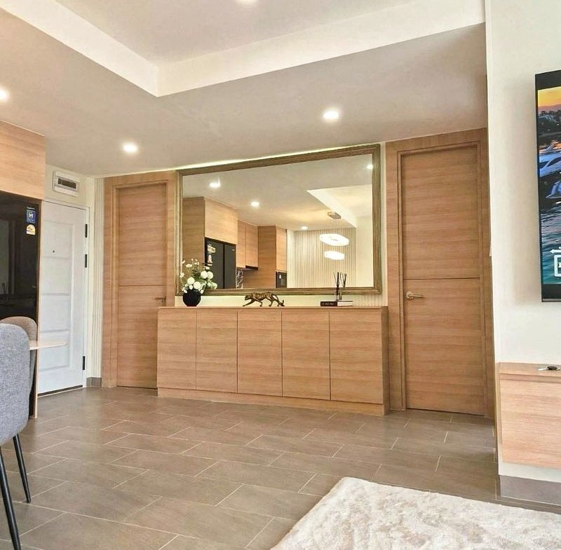 Entryway with light wood cabinets, a large mirror, and doorway; gray tile floor and a plush white rug nearby.