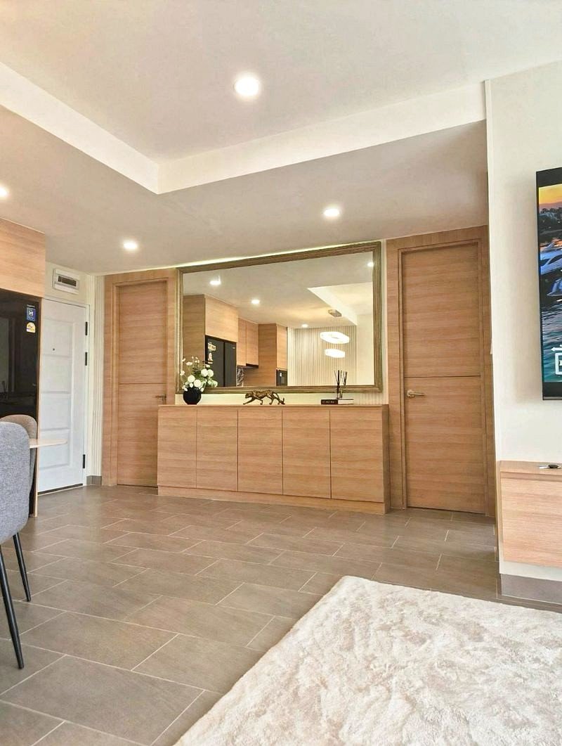 Entryway with light wood cabinets, a large mirror, and doorway; gray tile floor and a plush white rug nearby.