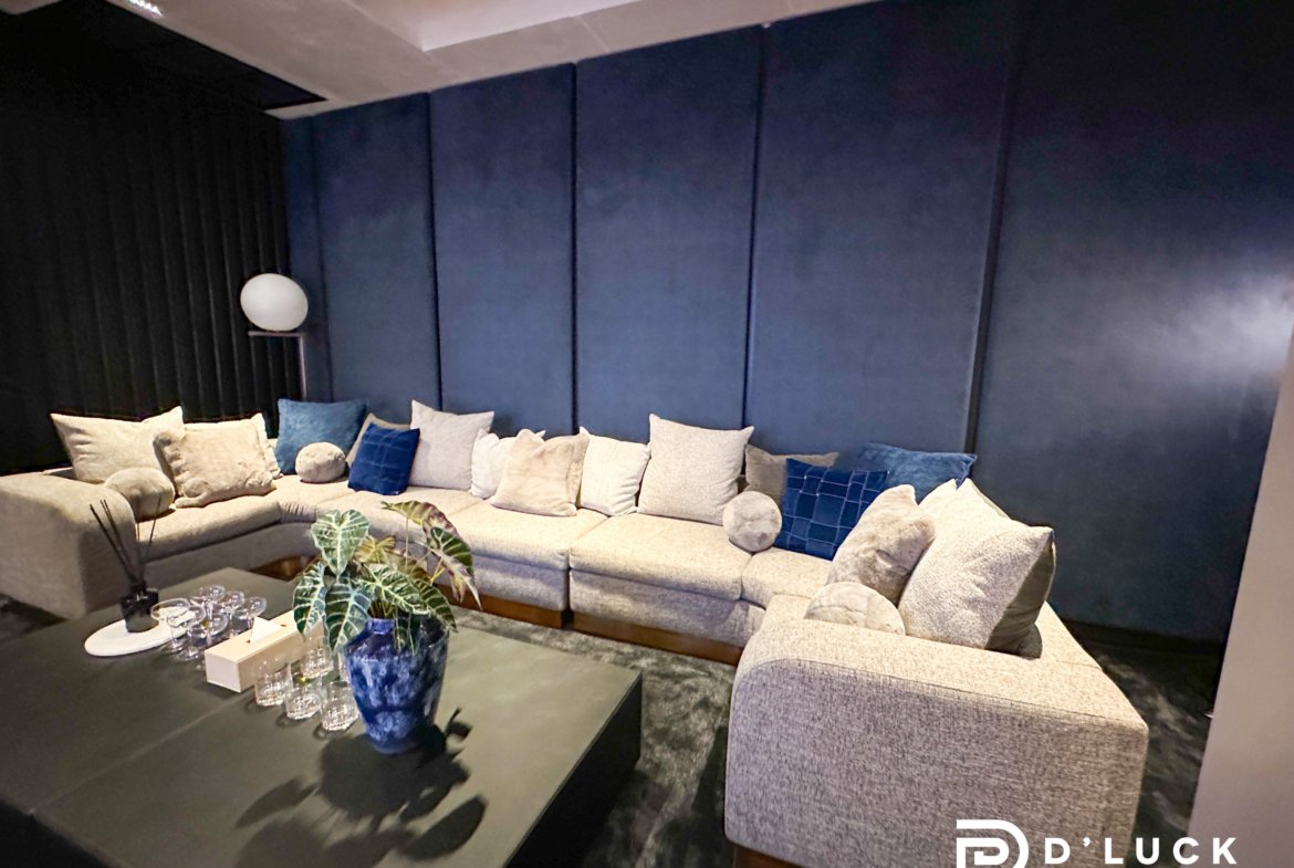 Cozy living room with a gray L-shaped sofa, cream and blue cushions, a dark blue padded wall, and a glass coffee table with a plant and glassware.
