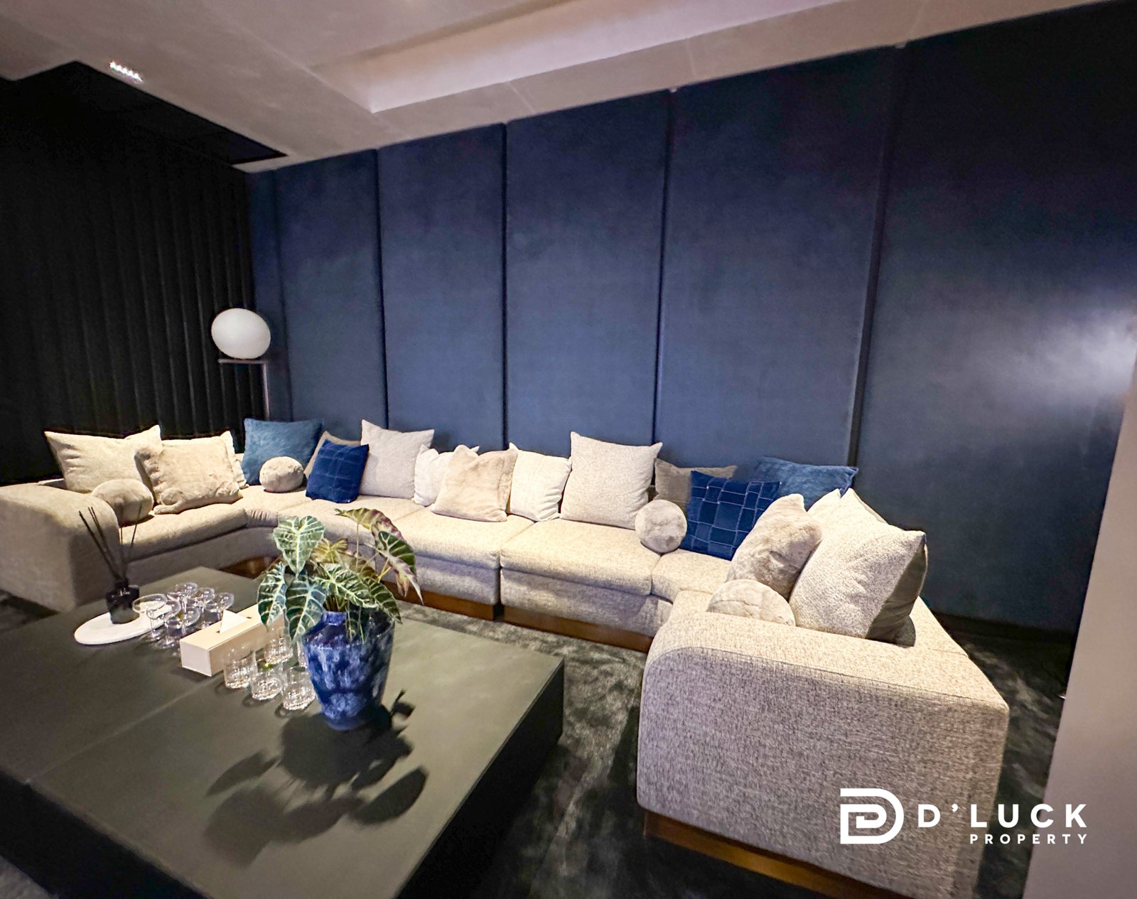 AW Common area 35 Cozy living room with a gray L-shaped sofa, cream and blue cushions, a dark blue padded wall, and a glass coffee table with a plant and glassware.