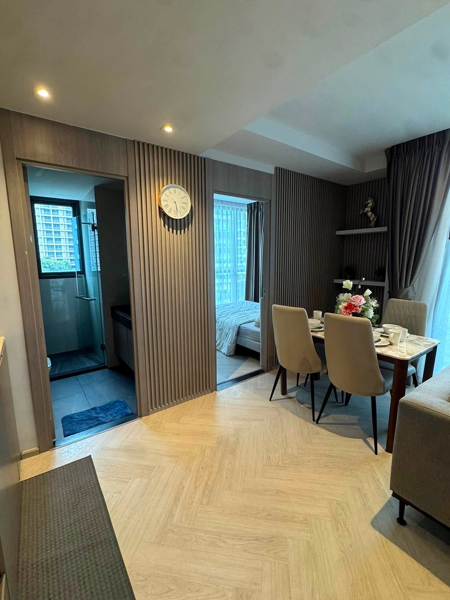 Andromeda_Pattaya_2Bed_Sale_Rent_Dlc631 Dining area in a modern apartment with beige chairs, a marble table set for a meal, and glass windows with dark drapes; flower centerpiece on the table.