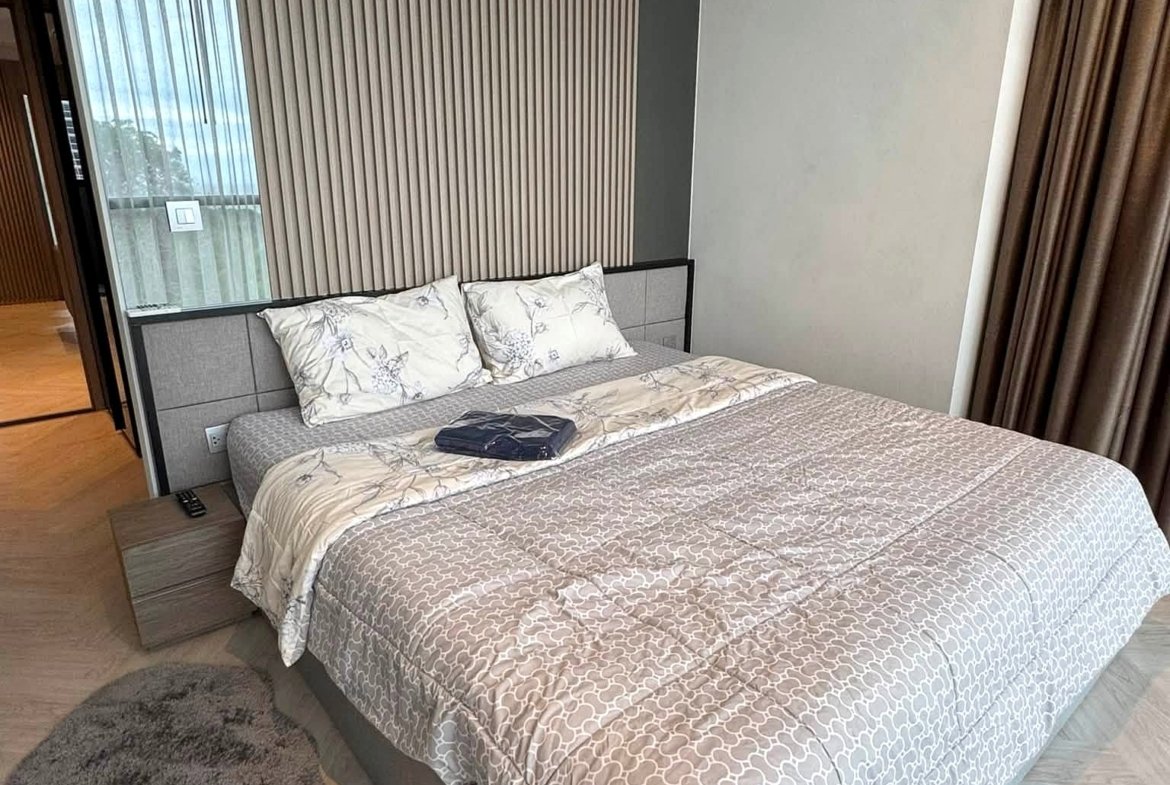 Modern hotel-style bedroom with a grey patterned duvet, decorative pillows, and a headboard against a vertical wood slat accent wall. Curtains frame a window on the right.