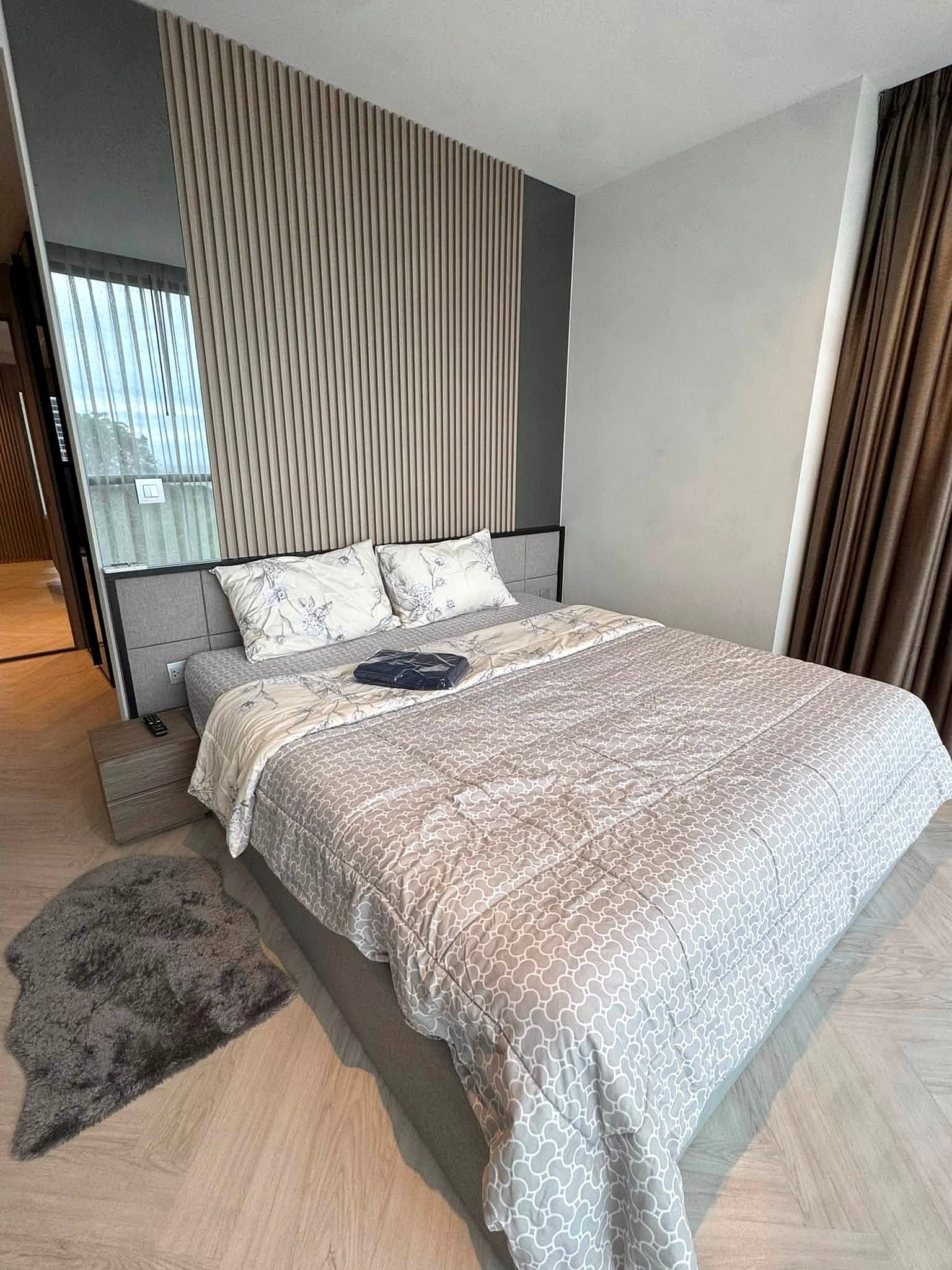Andromeda_Pattaya_2Bed_Sale_Rent_Dlc631_2 Modern hotel-style bedroom with a grey patterned duvet, decorative pillows, and a headboard against a vertical wood slat accent wall. Curtains frame a window on the right.
