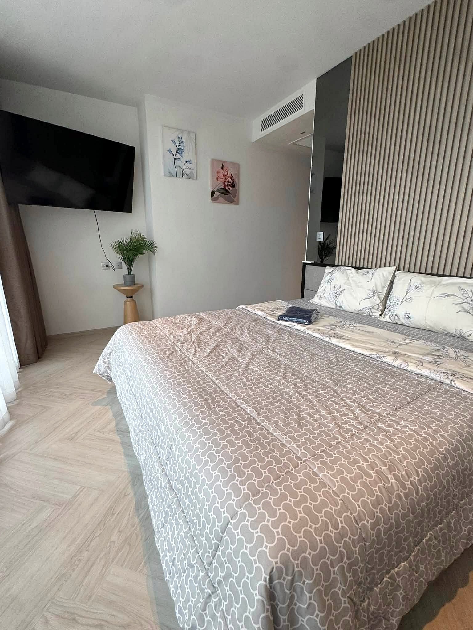 Andromeda_Pattaya_2Bed_Sale_Rent_Dlc631_3 A bedroom with a large bed covered in a gray geometric duvet, a slatted wooden headboard on the right, and a wall-mounted flat-screen TV on the left; a small plant on a wooden stand beside the TV and two framed botanical prints on the wall behind the bed.