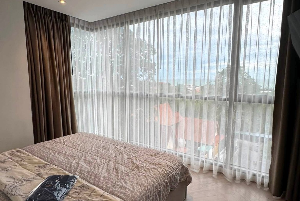 Hotel bedroom with two twin beds dressed in beige patterned quilts, facing large windows with sheer white curtains and brown drapes, soft natural light in the room.