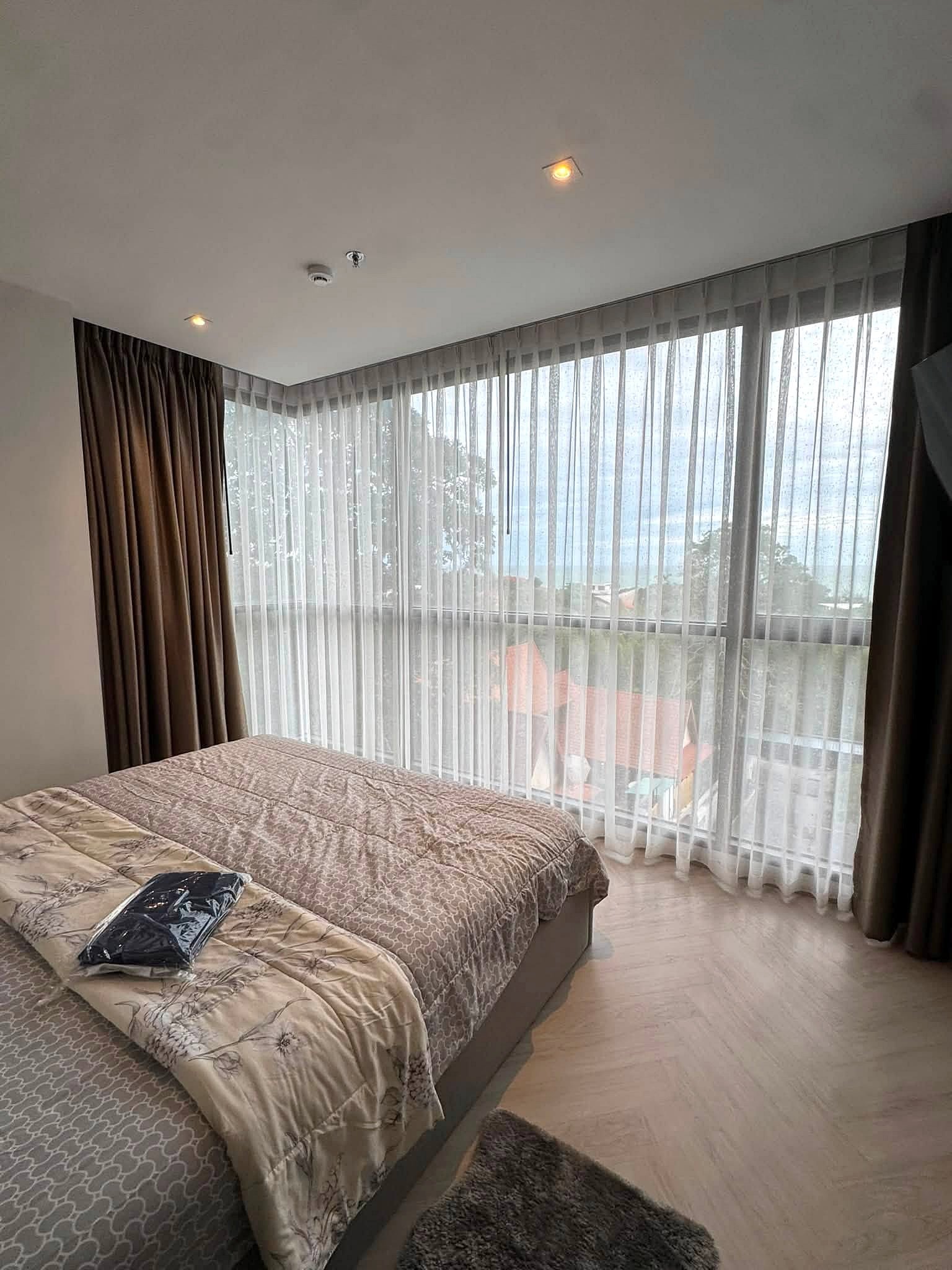 Andromeda_Pattaya_2Bed_Sale_Rent_Dlc631_4 Hotel bedroom with two twin beds dressed in beige patterned quilts, facing large windows with sheer white curtains and brown drapes, soft natural light in the room.