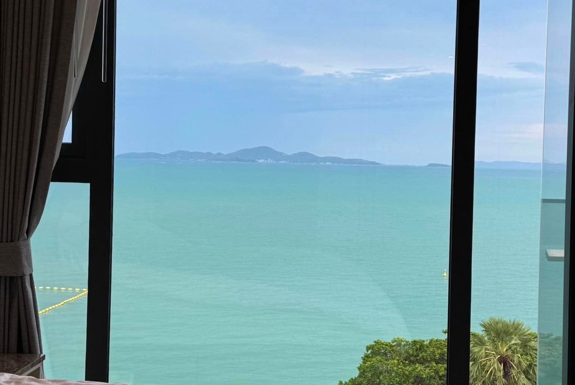 Bed with a white striped duvet in a room, large glass wall opens to the blue sea and distant islands.