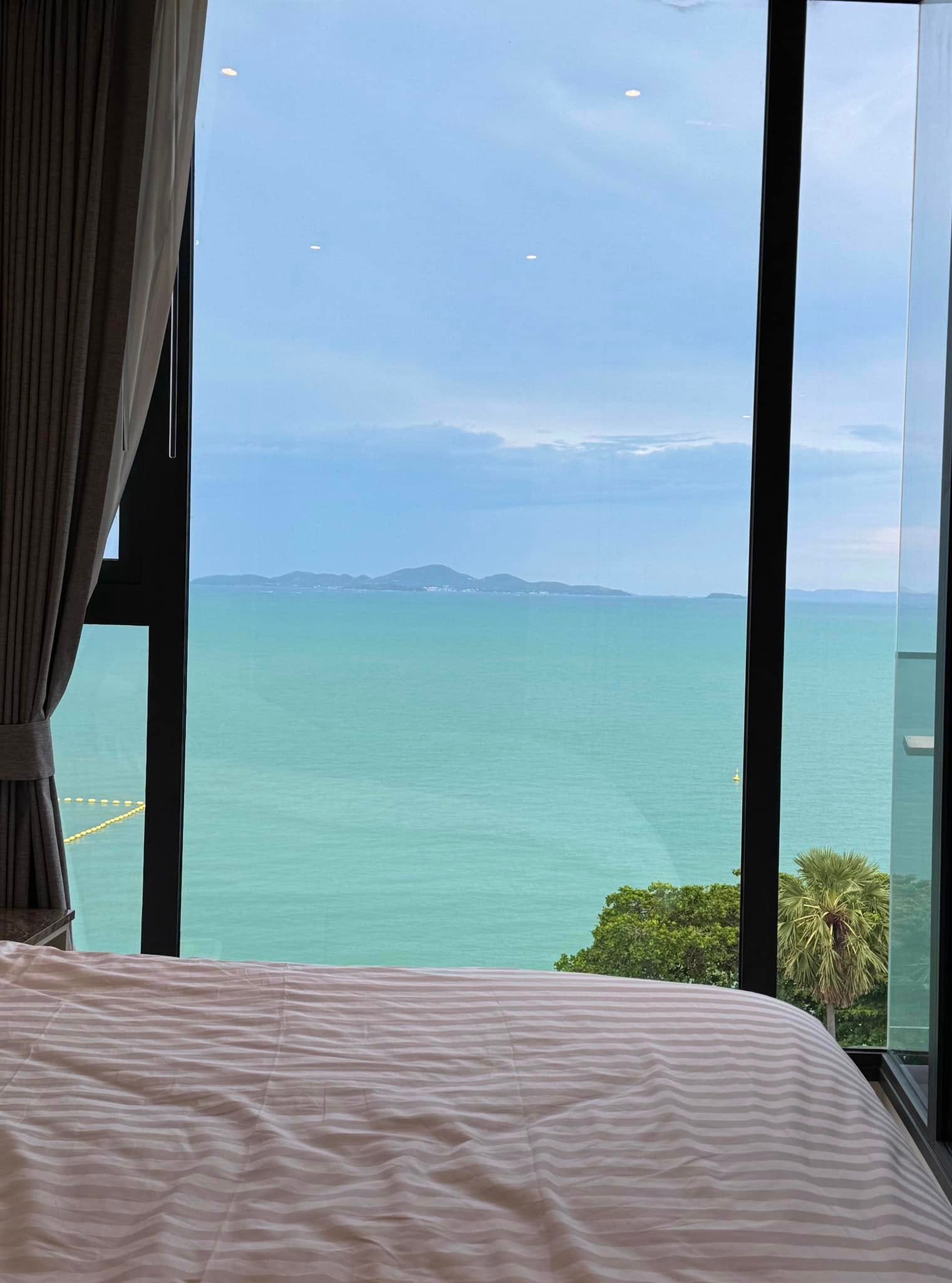 Arom_Wongamat_1005_10 Bed with a white striped duvet in a room, large glass wall opens to the blue sea and distant islands.