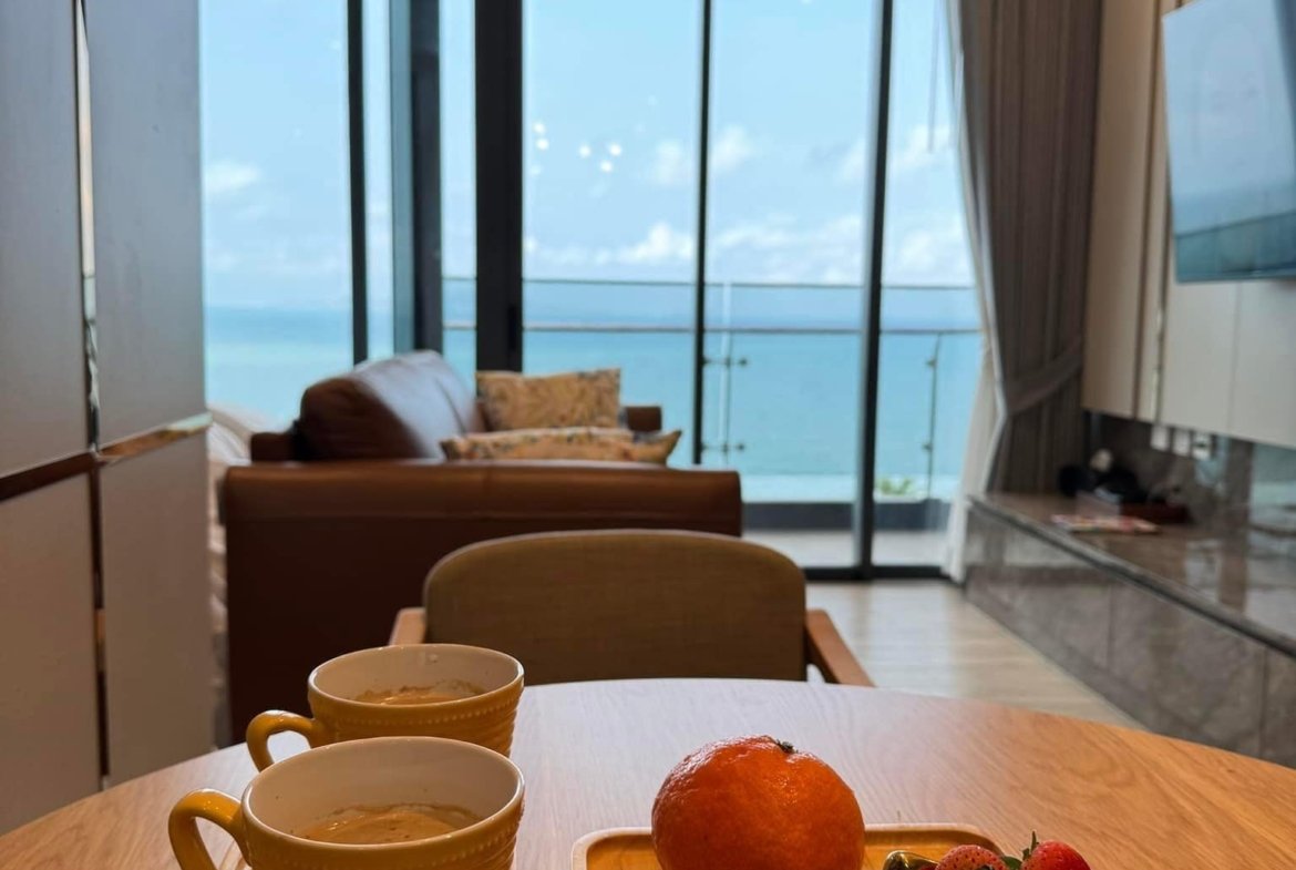 Cafe table with two coffee mugs, muffins, an orange, and strawberries on a wooden tray, with a sea-view through glass doors in the background.