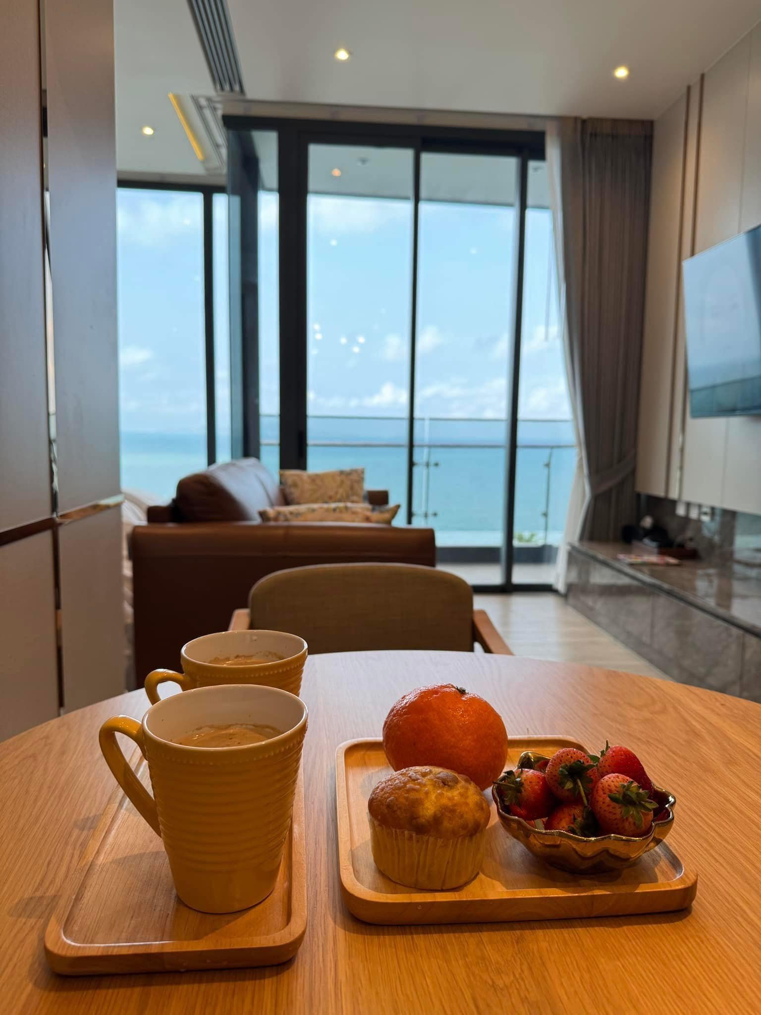 Arom_Wongamat_1005_2 Cafe table with two coffee mugs, muffins, an orange, and strawberries on a wooden tray, with a sea-view through glass doors in the background.