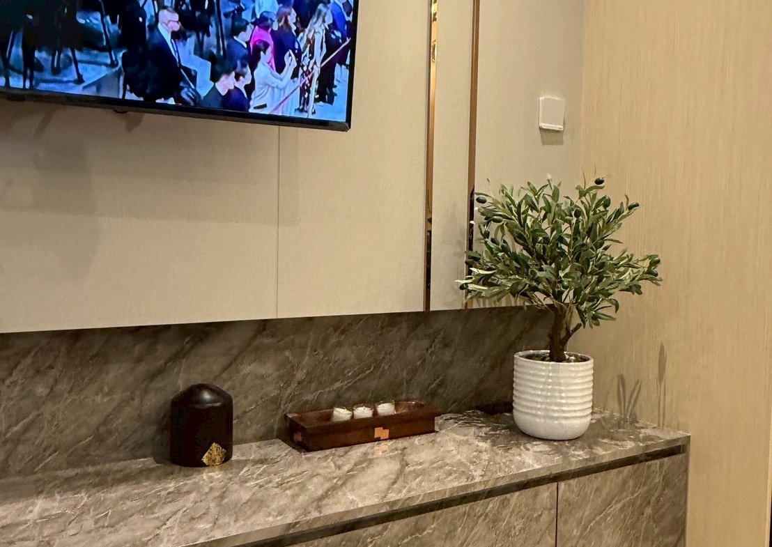 TV mounted on a beige wall above a gray marble console with a potted plant and a small dark candle on the left, tray of items in the center.