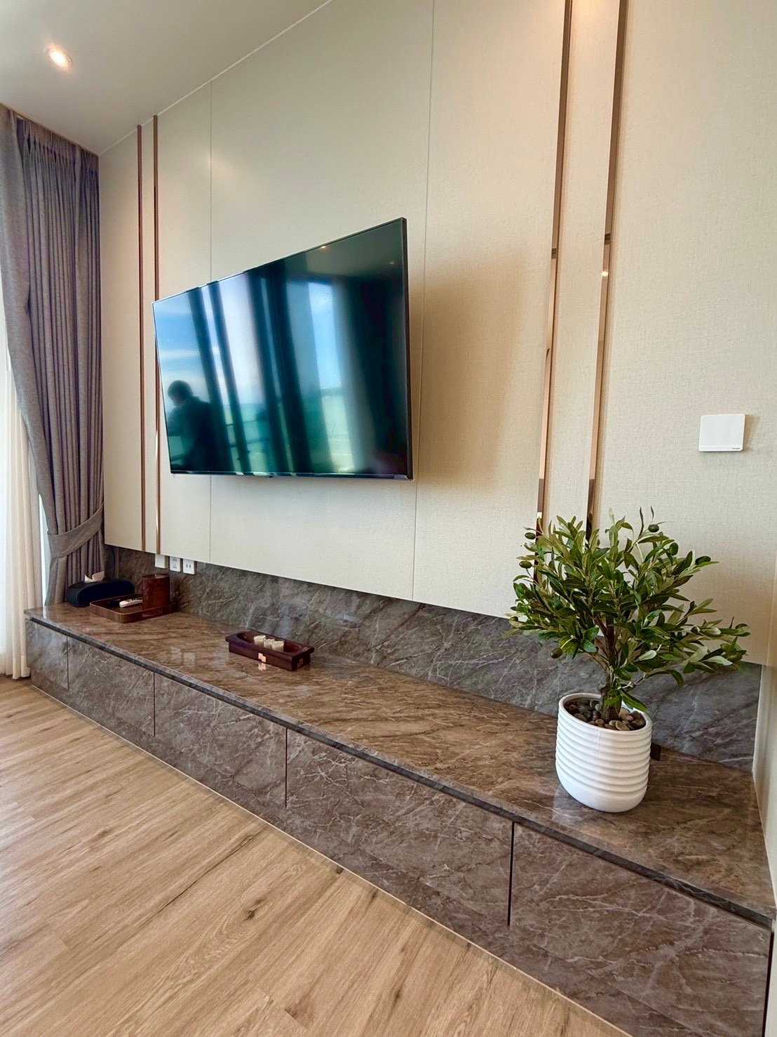 Arom_Wongamat_1005_4 Mounted flat-screen TV above a marble media console in a modern living room. The console spans the wall and holds small decorative items, with a leafy plant in a white pot on the right.