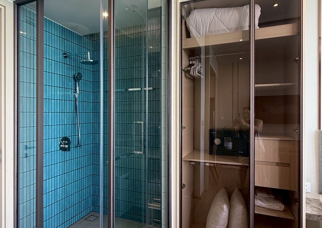 Modern bathroom with a glass-walled shower featuring blue tiled walls and a rainfall showerhead, beside a tall glass-front storage cabinet holding pillows and linens.
