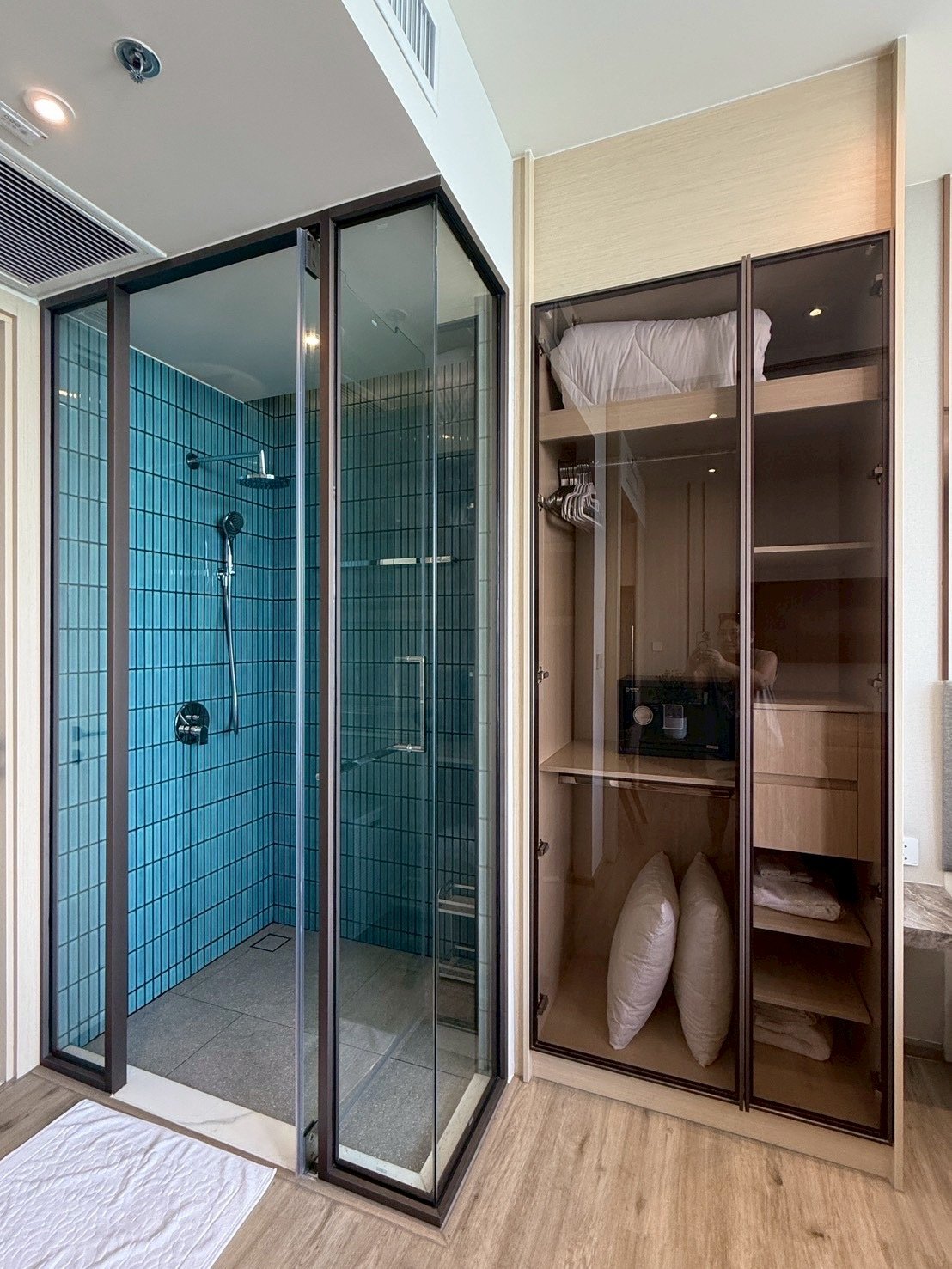 Arom_Wongamat_1005_8 Modern bathroom with a glass-walled shower featuring blue tiled walls and a rainfall showerhead, beside a tall glass-front storage cabinet holding pillows and linens.