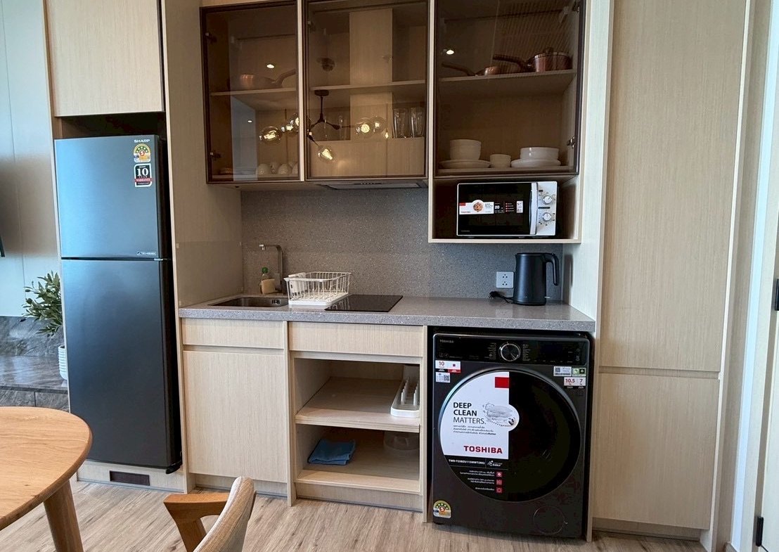 Compact kitchen with a tall refrigerator, glass-front upper cabinets, microwave, kettle, washing machine, and a sink with dish rack.