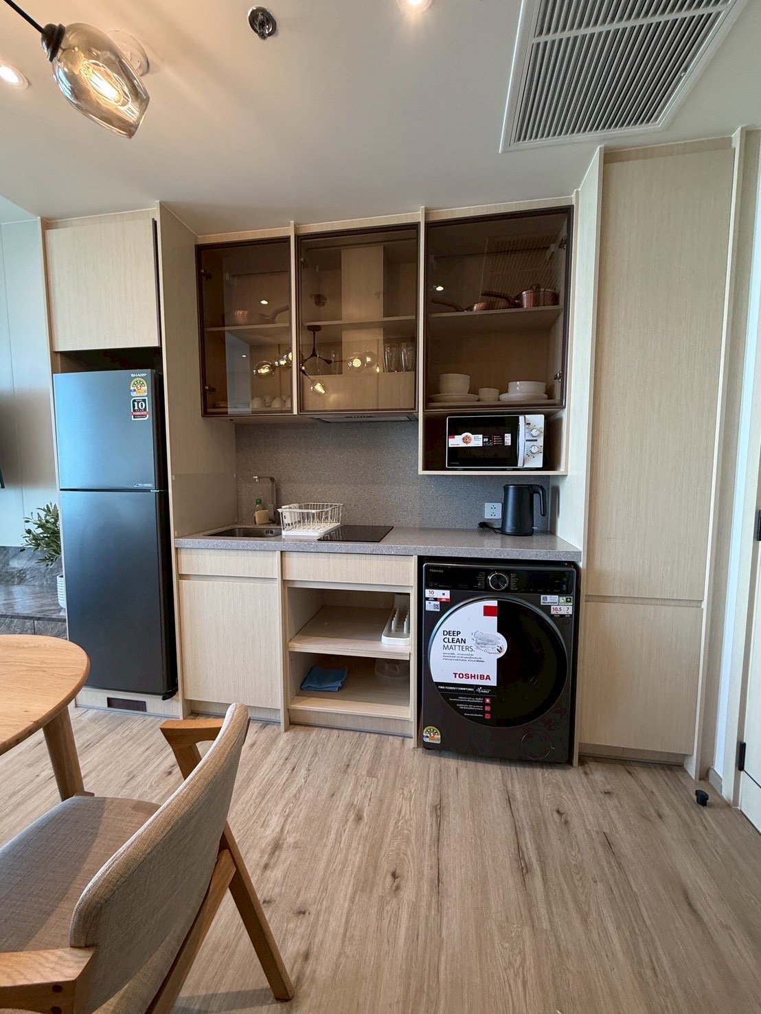 Arom_Wongamat_1005_9 Compact kitchen with a tall refrigerator, glass-front upper cabinets, microwave, kettle, washing machine, and a sink with dish rack.