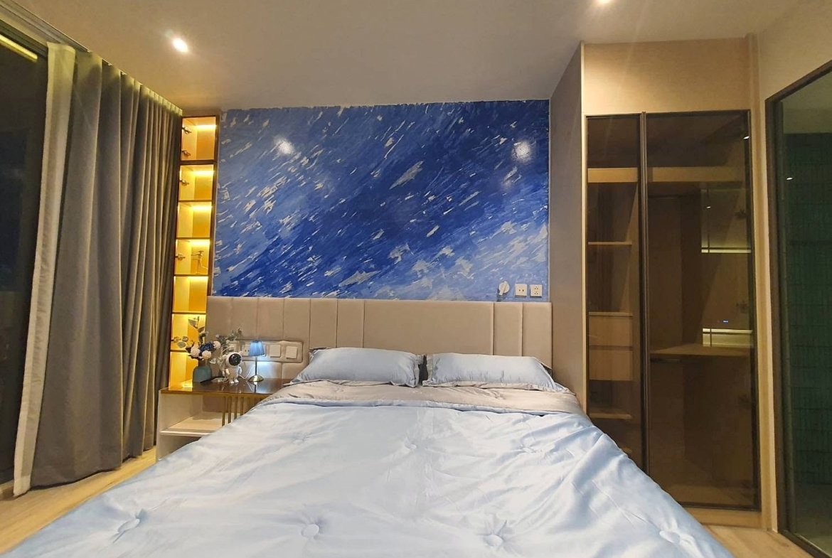 Bedroom with a blue abstract accent wall behind a padded beige headboard; light blue bedding and pillows, side table with flowers and lamp on the left.
