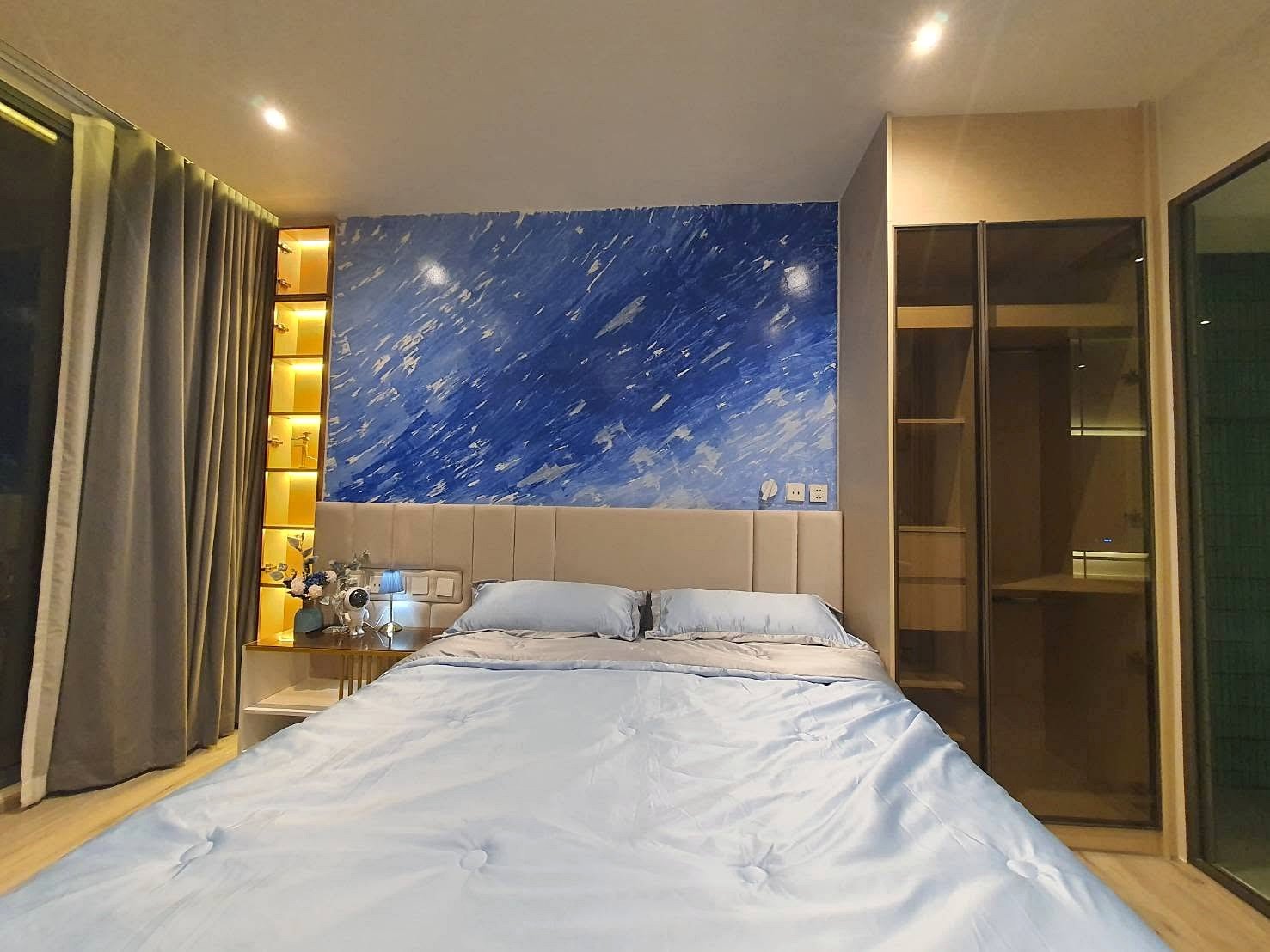 Arom_Wongamat_1bed_Rent_Rdl334_10 Bedroom with a blue abstract accent wall behind a padded beige headboard; light blue bedding and pillows, side table with flowers and lamp on the left.