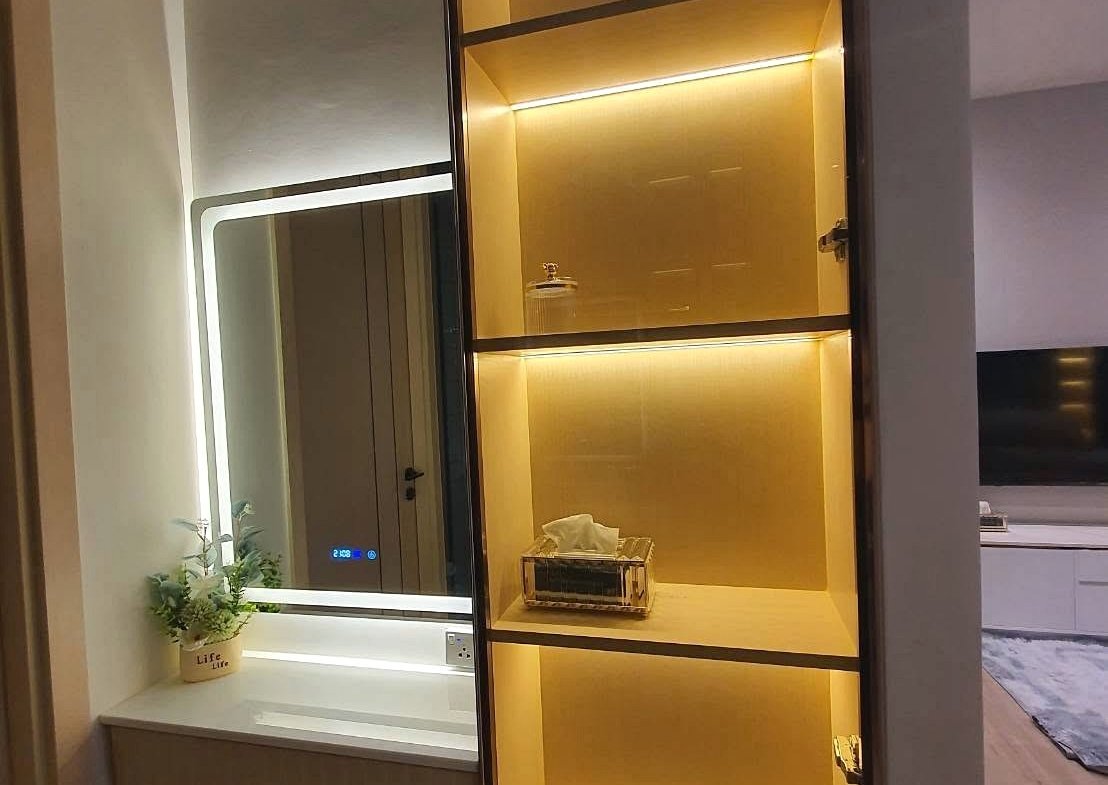 Tall, illuminated shelving unit with warm yellow lighting in a modern living space, adjacent to a vanity desk with a backlit mirror and a small potted plant nearby
