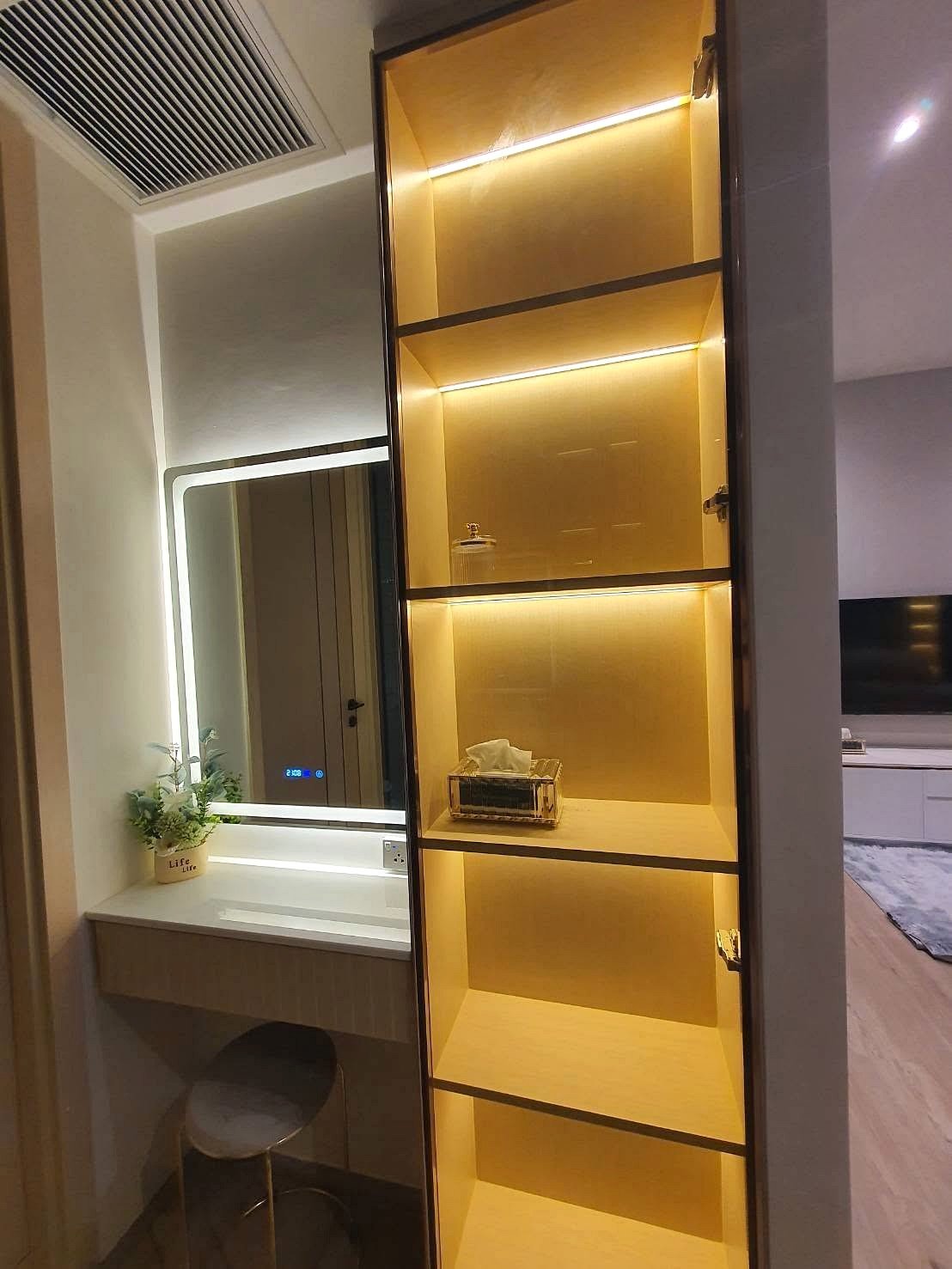 Arom_Wongamat_1bed_Rent_Rdl334_12 Tall, illuminated shelving unit with warm yellow lighting in a modern living space, adjacent to a vanity desk with a backlit mirror and a small potted plant nearby