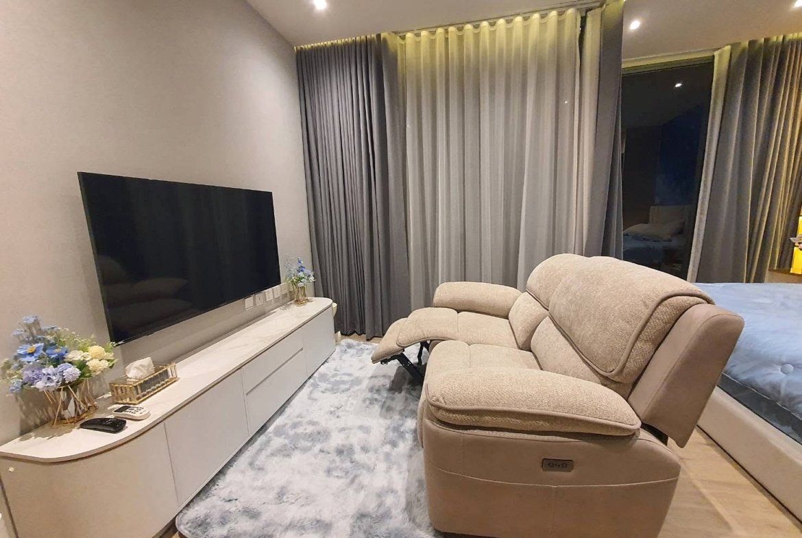 Living room with a large flat-screen TV on a white media console, beige reclining chairs, and blue floral arrangements by the curtains.
