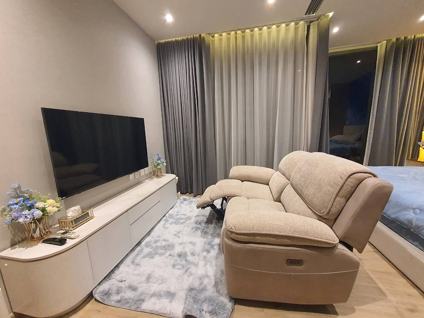 Arom_Wongamat_1bed_Rent_Rdl334_13 Living room with a large flat-screen TV on a white media console, beige reclining chairs, and blue floral arrangements by the curtains.