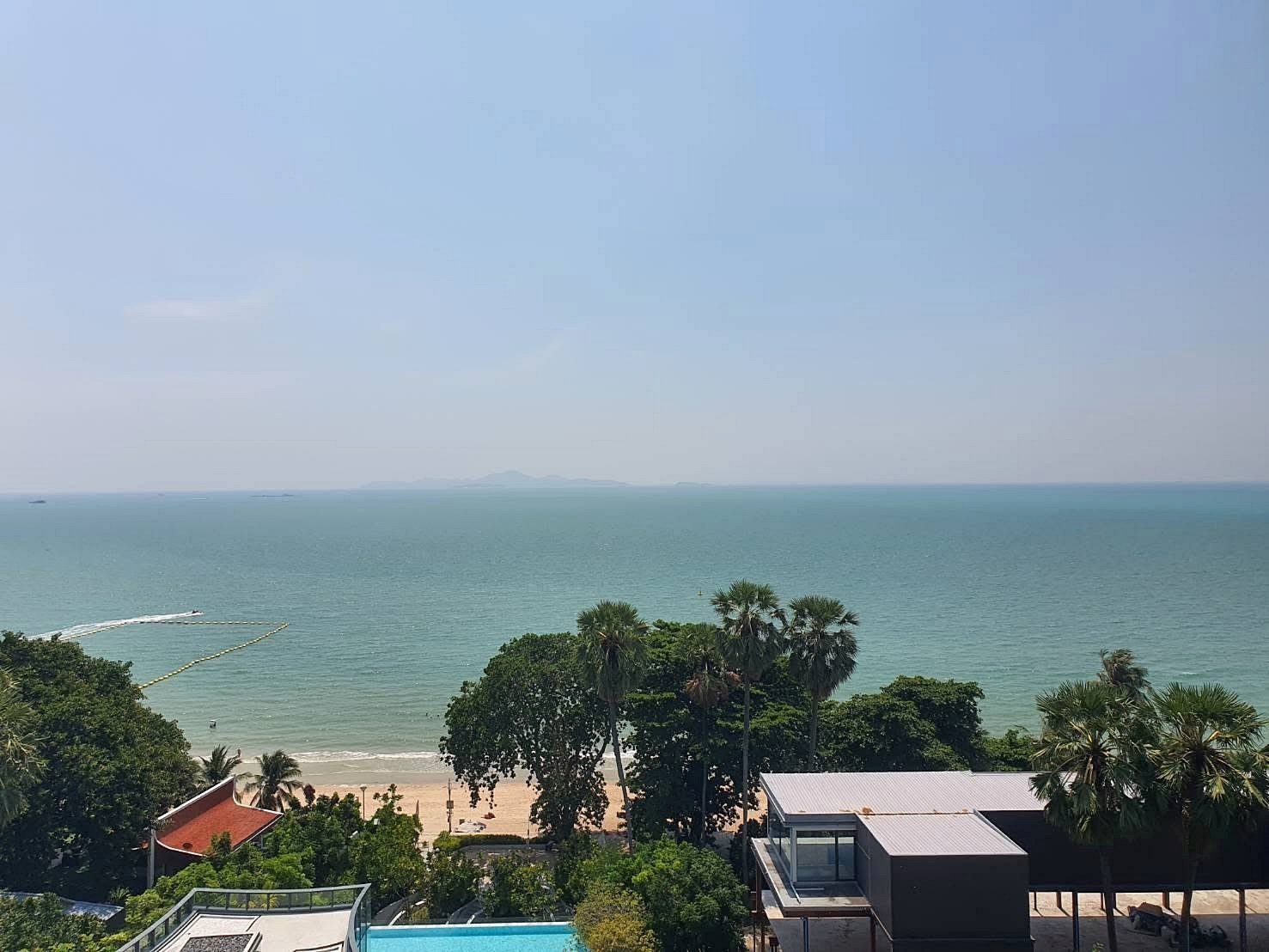 Arom_Wongamat_1bed_Rent_Rdl334_14 Coastal beachfront with palm trees, a sandy shore, and calm turquoise sea stretching to the horizon under a clear blue sky.