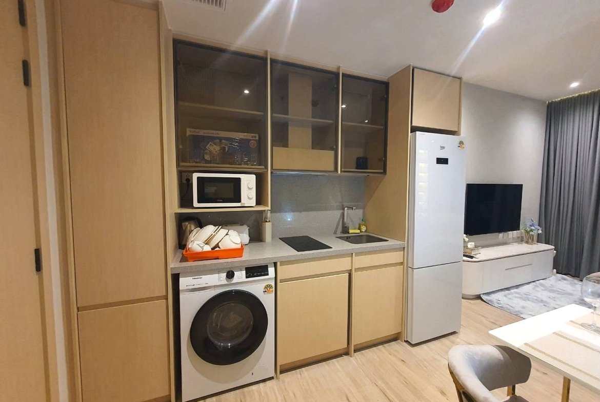 Modern kitchenette with light wood cabinets, upper glass-front cupboards, microwave, washing machine, and a small sink next to an induction hob; a tall fridge stands nearby as a living room with a TV and sofa is visible.