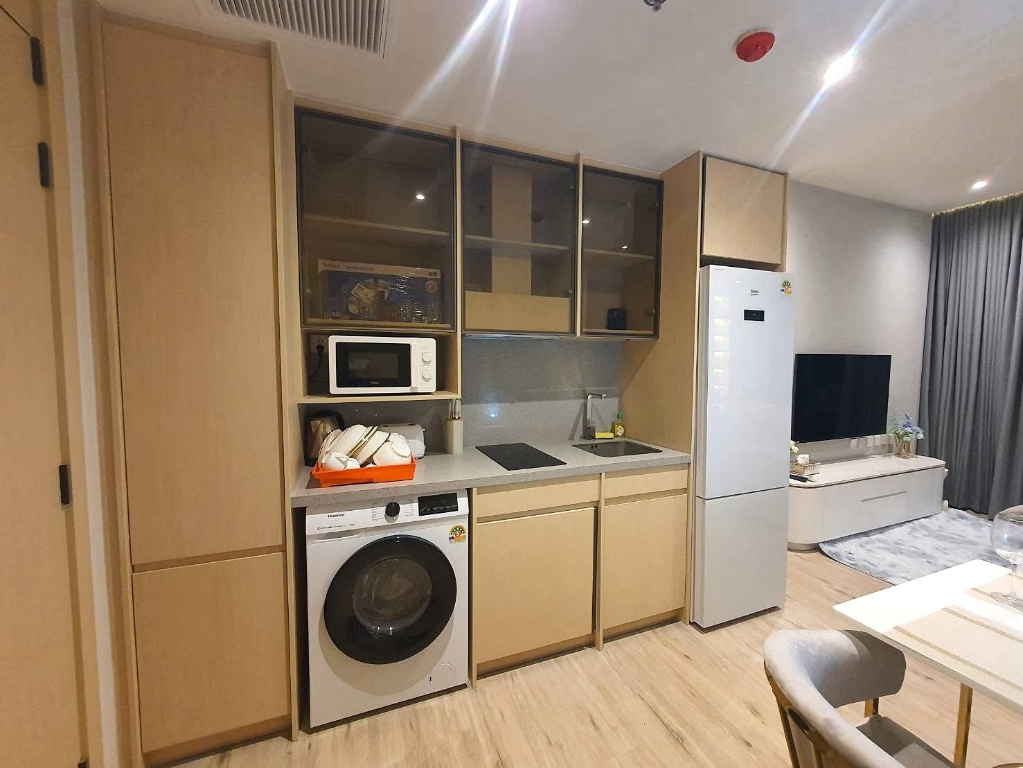 Arom_Wongamat_1bed_Rent_Rdl334_3 Modern kitchenette with light wood cabinets, upper glass-front cupboards, microwave, washing machine, and a small sink next to an induction hob; a tall fridge stands nearby as a living room with a TV and sofa is visible.