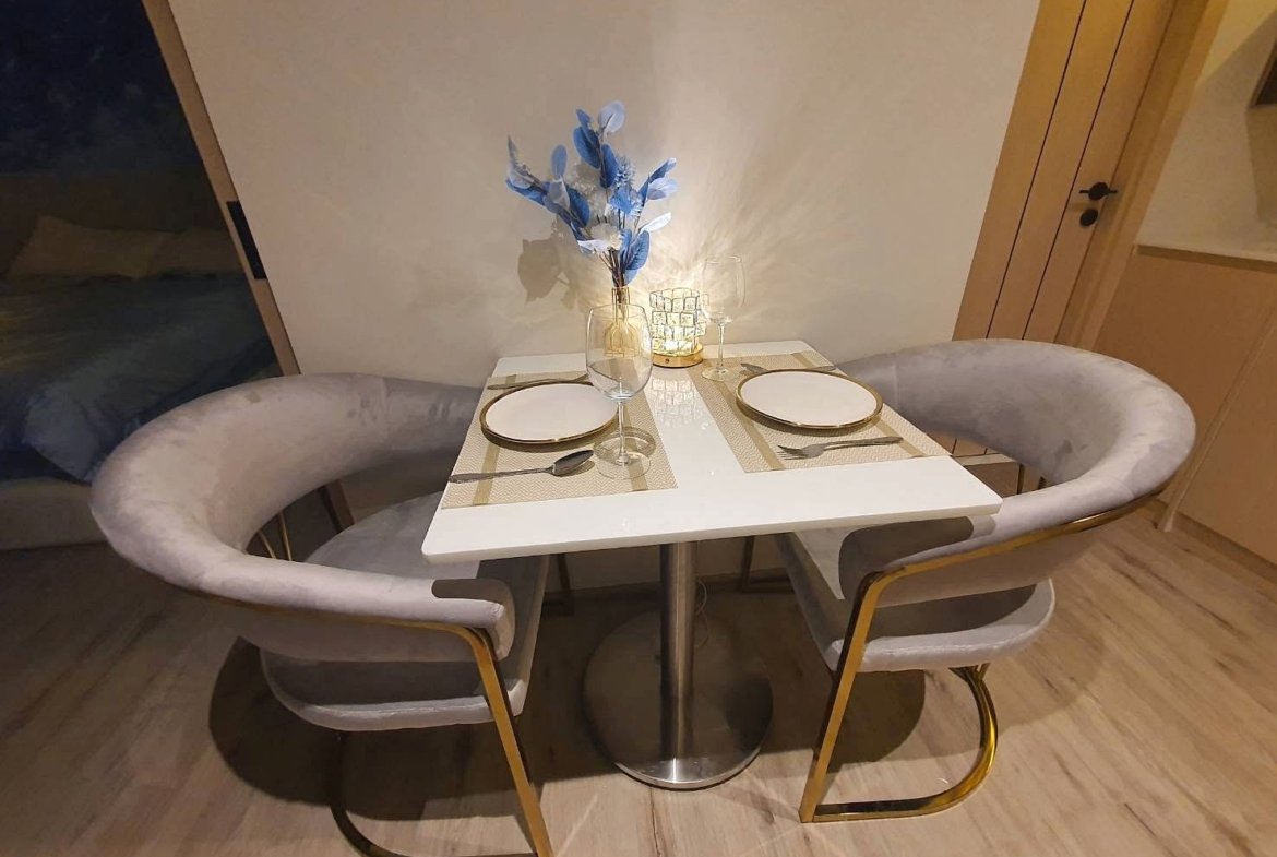 Square dining table set for two with beige placemats, white plates, and wine glasses, centerpiece of blue leaves and a lit candle.