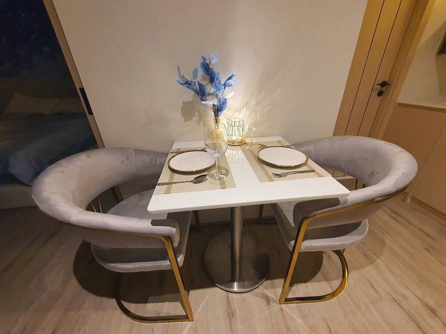 Arom_Wongamat_1bed_Rent_Rdl334_4 Square dining table set for two with beige placemats, white plates, and wine glasses, centerpiece of blue leaves and a lit candle.