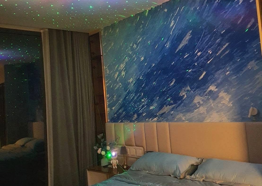 Bedroom with blue wave mural, starry ceiling projection, and a large bed with blue sheets; nightstand and lamp at the headboard.
