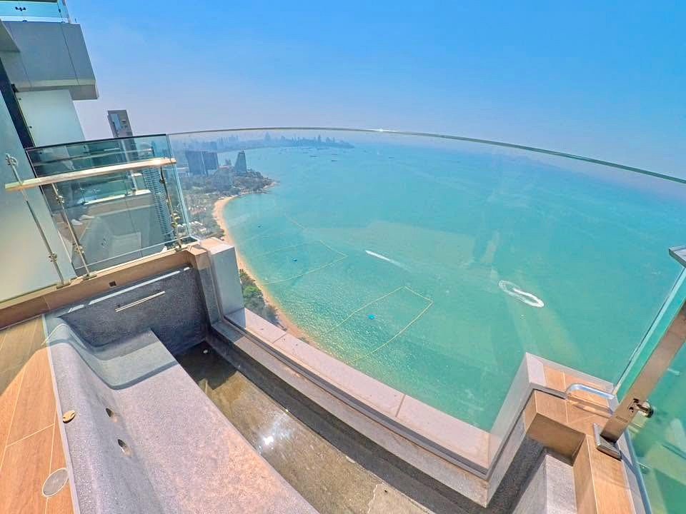 Balcony with glass railing overlooking turquoise sea and a sandy beach on a sunny day, city skyline in the distance.