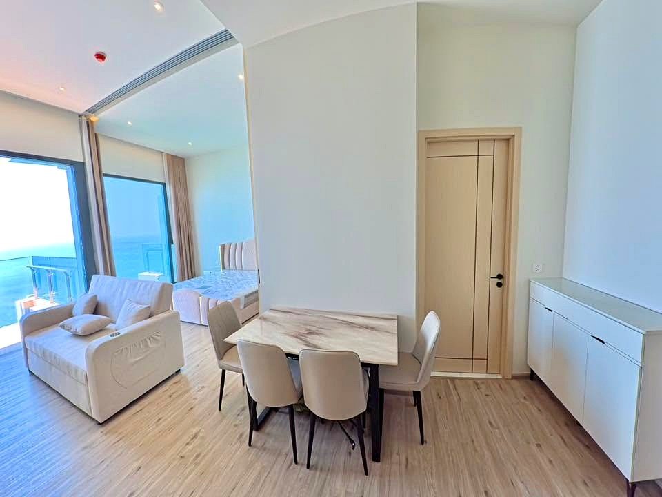 Open-plan living and dining area with a marble dining table, white chairs, and a sofa, with a sea-view balcony visible through large windows.